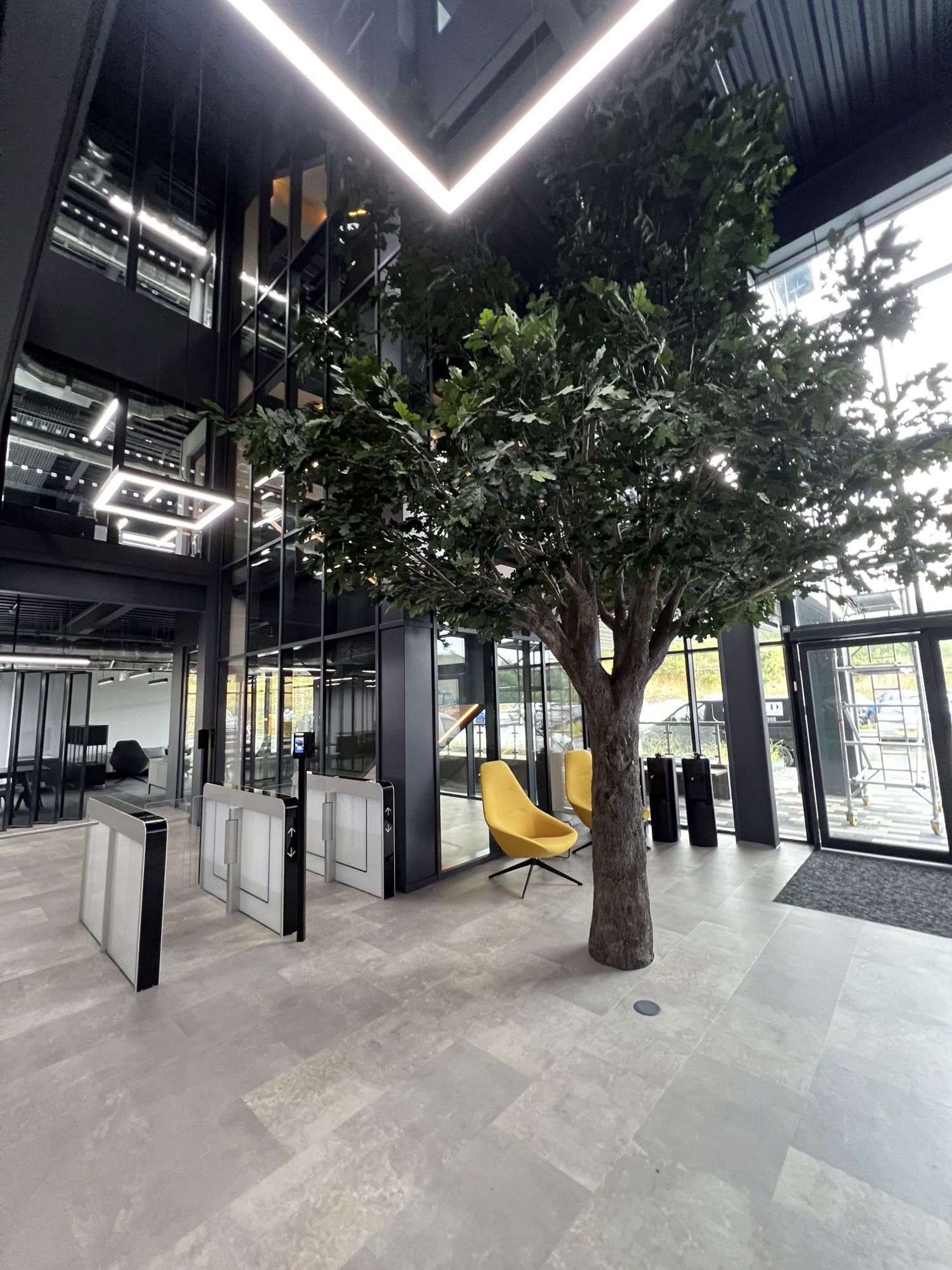 Modernes Bürointerieur mit einem großen künstlichen Baum, gelben Sitzmöbeln, Glaswänden und Sicherheitsdrehkreuzen.